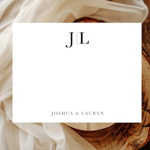 May include: A white card with the initials "JL" in black, with the names "Joshua & Lauren" below. The card is set against a backdrop of draped fabric and a wooden surface.