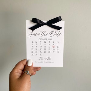 K&ouml;nnte beinhalten: Eine wei&szlig;e Save-the-Date-Karte mit einem schwarzen Band, das darum gebunden ist. Die Karte hat einen Kalender mit dem hervorgehobenen Datum 13. Oktober 2023. Der Text "Save the Date" ist oben auf der Karte in Kursivschrift geschrieben. Die Namen "Joni & Alex" und der Ort "Ft. Lauderdale, Florida" sind unten auf der Karte gedruckt.
