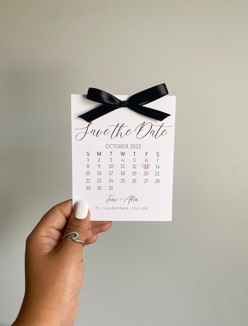 K&ouml;nnte beinhalten: Eine wei&szlig;e Save-the-Date-Karte mit einem schwarzen Band, das darum gebunden ist. Die Karte hat einen Kalender mit dem hervorgehobenen Datum 13. Oktober 2023. Der Text "Save the Date" ist oben auf der Karte in Kursivschrift geschrieben. Die Namen "Joni & Alex" und der Ort "Ft. Lauderdale, Florida" sind unten auf der Karte gedruckt.