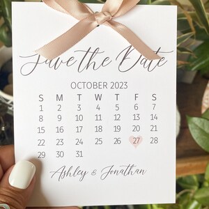 K&ouml;nnte beinhalten: Save the Date Karte mit einem Kalender f&uuml;r Oktober 2023. Der 27. ist mit einem rosa Herz hervorgehoben. Die Karte ist mit einem beigefarbenen Band verziert, das zu einer Schleife gebunden ist. Der Text unten lautet "Ashley & Jonathan".