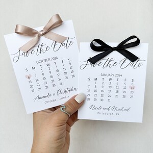 K&ouml;nnte beinhalten: Zwei Save-the-Date-Karten mit Kalendern f&uuml;r Oktober und Januar. Die Oktoberkarte hat eine hellbraune Schleife und die Januar-Karte eine schwarze Schleife. Beide Karten haben ein kleines rosa Herz, das das Datum markiert. Die Oktoberkarte lautet "Amanda & Christopher" und "Chicago, Illinois". Die Januar-Karte lautet "Nicole und Michael" und "Pittsburgh, PA".