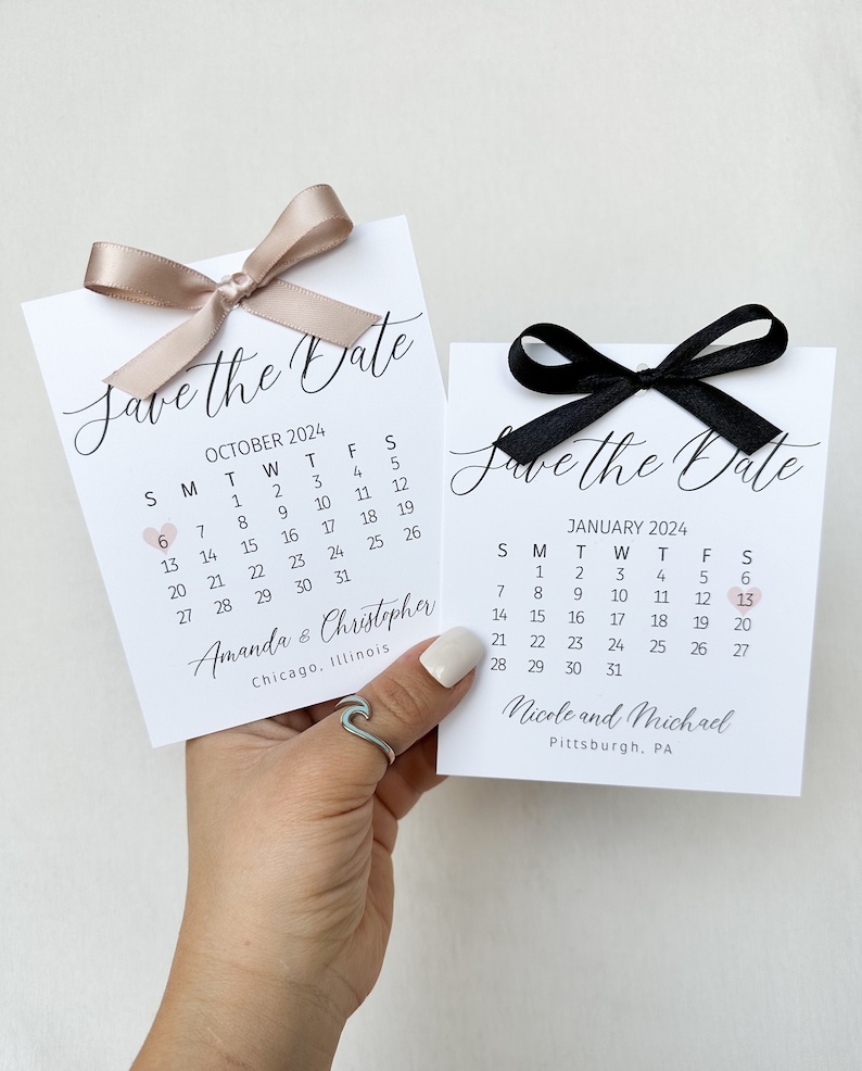 K&ouml;nnte beinhalten: Zwei Save-the-Date-Karten mit Kalendern f&uuml;r Oktober und Januar. Die Oktoberkarte hat eine hellbraune Schleife und die Januar-Karte eine schwarze Schleife. Beide Karten haben ein kleines rosa Herz, das das Datum markiert. Die Oktoberkarte lautet "Amanda & Christopher" und "Chicago, Illinois". Die Januar-Karte lautet "Nicole und Michael" und "Pittsburgh, PA".