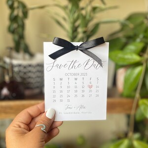 K&ouml;nnte beinhalten: Save the Date Karte mit einer schwarzen Schleife und einem Kalender mit einem Herz, das den 13. Oktober 2023 markiert. Die Karte lautet "Save the Date" und "Joni + Alex" mit dem Ort "Ft. Lauderdale, Florida".
