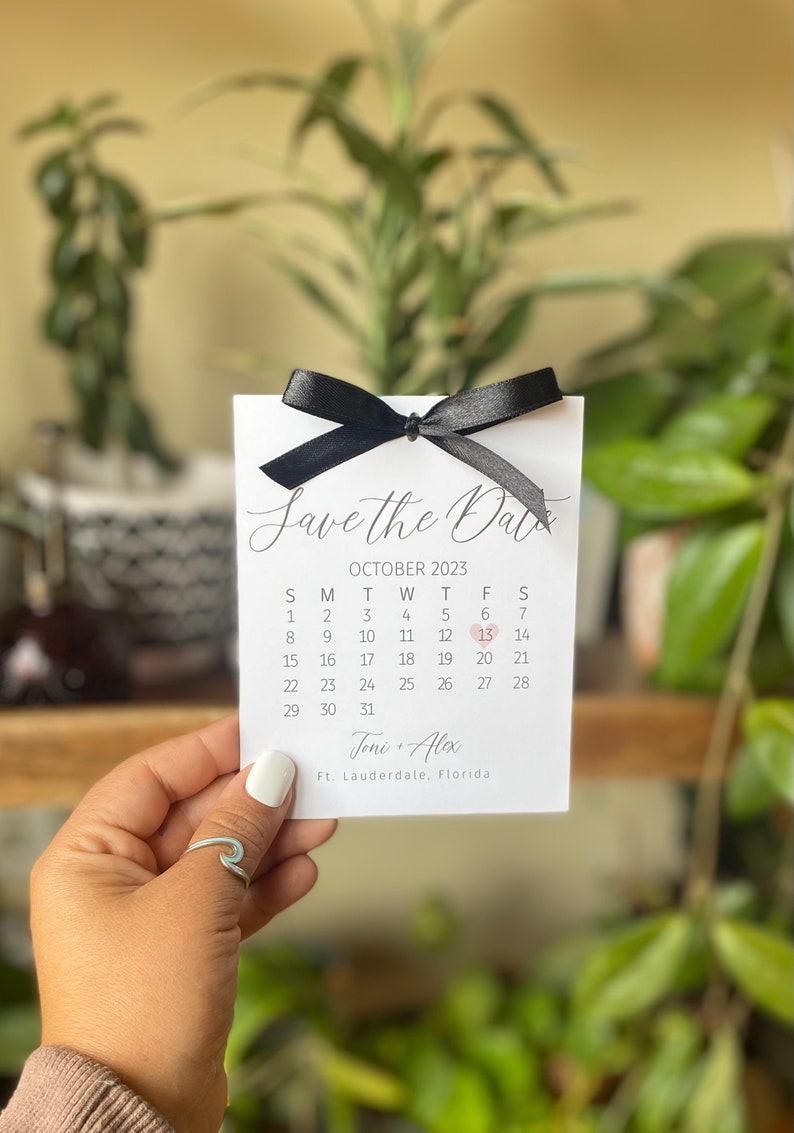 K&ouml;nnte beinhalten: Save the Date Karte mit einer schwarzen Schleife und einem Kalender mit einem Herz, das den 13. Oktober 2023 markiert. Die Karte lautet "Save the Date" und "Joni + Alex" mit dem Ort "Ft. Lauderdale, Florida".