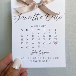 K&ouml;nnte beinhalten: Save-the-Date-Karte mit einem Kalender f&uuml;r August 2023, wobei der 26. in Rosa eingekreist ist. Der Text "Isla Grace You're going to be our flower girl!" ist unter dem Kalender gedruckt.