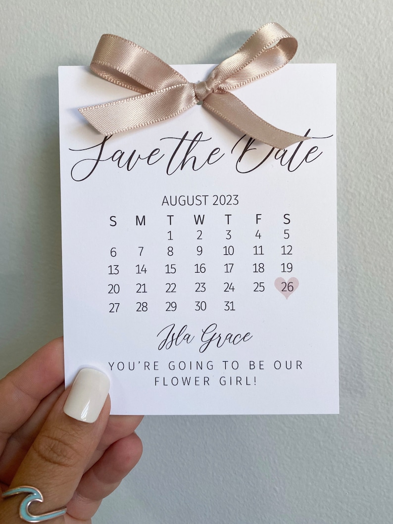 K&ouml;nnte beinhalten: Save-the-Date-Karte mit einem Kalender f&uuml;r August 2023, wobei der 26. in Rosa eingekreist ist. Der Text "Isla Grace You're going to be our flower girl!" ist unter dem Kalender gedruckt.