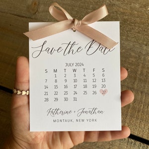 K&ouml;nnte beinhalten: Save the Date Karte mit wei&szlig;em Hintergrund und einem rosa Band. Die Karte zeigt einen Kalender mit dem Datum 27. Juli, das mit einem rosa Herz hervorgehoben ist. Der Text "Katherine + Jonathan" und "Montauk, New York" ist unter dem Kalender gedruckt.
