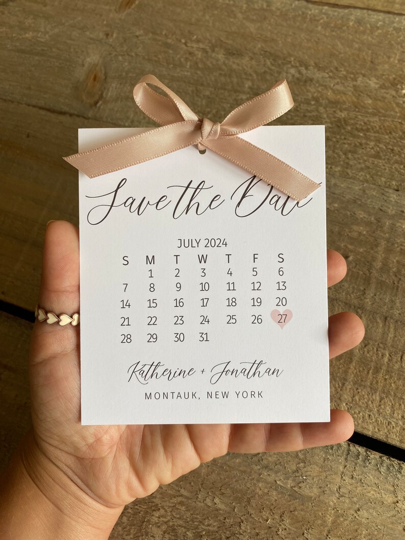 K&ouml;nnte beinhalten: Save the Date Karte mit wei&szlig;em Hintergrund und einem rosa Band. Die Karte zeigt einen Kalender mit dem Datum 27. Juli, das mit einem rosa Herz hervorgehoben ist. Der Text "Katherine + Jonathan" und "Montauk, New York" ist unter dem Kalender gedruckt.
