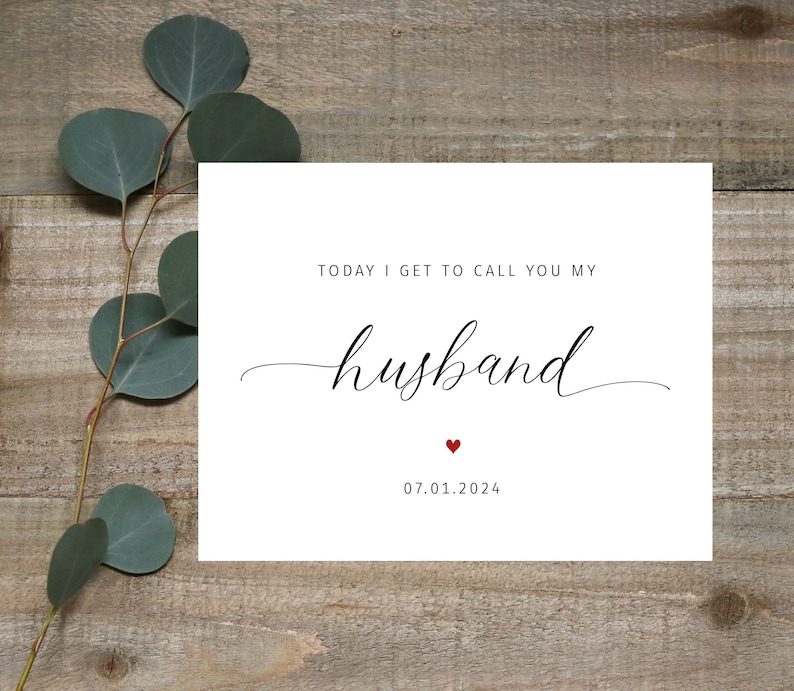 May include: A white card with the text "TODAY I GET TO CALL YOU MY husband" in black script. A small red heart is below the word "husband", and the date "07.01.2024" is printed below the heart. The card is on a wooden surface.