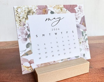 May include: A floral desktop calendar with a wooden stand. The calendar features a watercolor floral design in shades of pink, beige, and green. The month of May 2024 is displayed on the calendar.