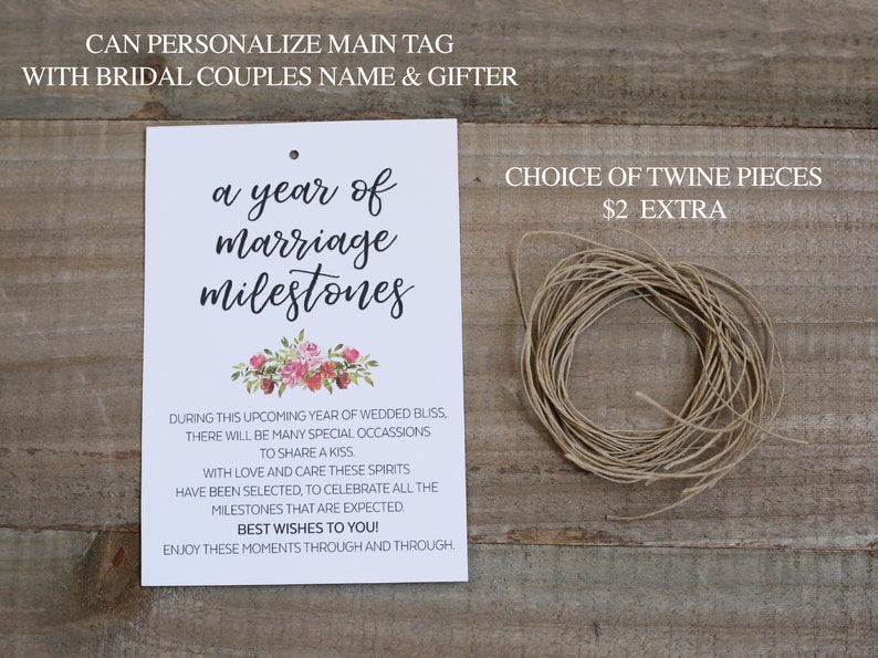 May include: A white card with the text "a year of marriage milestones" in black script. The card has a floral design with pink and white flowers. The text "CHOICE OF TWINE PIECES &pound;2 EXTRA" is in black text on a white background. A roll of twine is shown on a wooden surface.
