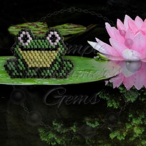 May include: A beaded frog figurine in shades of green, yellow, and black sits on a lily pad. A pink and white water lily blooms nearby, reflected in the dark water. The scene evokes a tranquil pond setting.