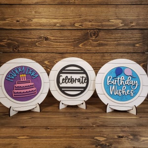 May include: Three white wooden round signs with a white background and a black stand. The signs have a circle in the center with different designs. The first sign says "Happy Day" with a purple cake and candles. The second sign says "Celebrate" with black stripes. The third sign says "Birthday Wishes" with blue and pink balloons.