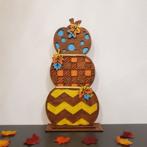 May include: A wooden fall pumpkin stack decoration with three pumpkins. The bottom pumpkin is brown with yellow zigzags, the middle pumpkin is brown with orange and white checkered pattern, and the top pumpkin is brown with blue polka dots. Each pumpkin has a small orange and yellow leaf on top.