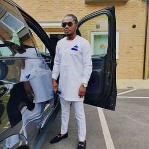 May include: A man wearing a white long-sleeved shirt and matching pants, with a blue and white design on the chest. He is wearing black sunglasses and black loafers. He is standing next to a black car with the door open.
