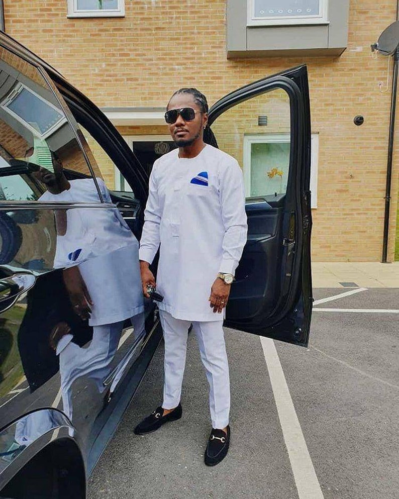 May include: A man wearing a white long-sleeved shirt and matching pants, with a blue and white design on the chest. He is wearing black sunglasses and black loafers. He is standing next to a black car with the door open.