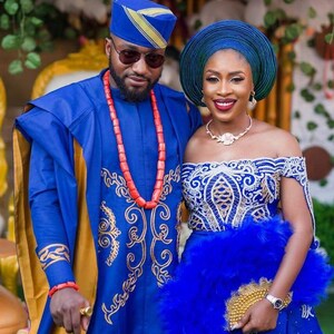 May include: A couple in traditional Nigerian attire. The man is wearing a blue and gold robe with a red beaded necklace. The woman is wearing a blue and white lace dress with a blue feathered fan. They are both smiling.