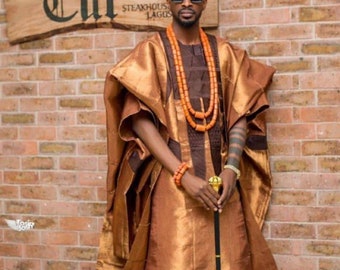 May include: A man wearing a gold and brown traditional African outfit with a matching head wrap. He is wearing sunglasses and a beaded necklace. He is holding a walking stick with a gold top.