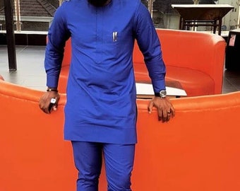 Puede incluir: Un hombre con una camisa de manga larga azul real y pantalones a juego. Está de pie frente a un sofá naranja y lleva gafas de sol.