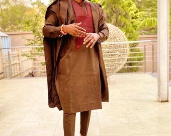 May include: A man wearing a brown traditional outfit with a maroon vest and a patterned hat. He is standing on a tiled floor.
