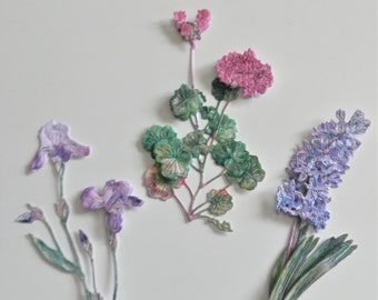 以下が含まれることがあります： 異なる色とスタイルの紙の花4つ。紫のアイリス、ピンクのゼラニウム、紫のヒアシンス、ピンクの花と緑の葉。