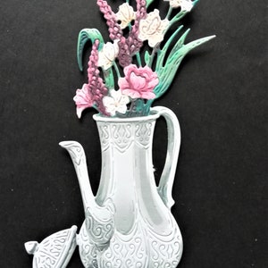 May include: A white ceramic teapot with an ornate design and a lid. The teapot is filled with a bouquet of pink, white, and green flowers.