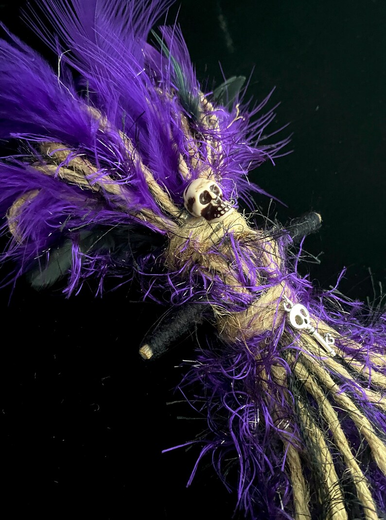 Op de afbeelding: Een handgemaakt decoratief item met paarse veren, beige touw en zwarte accenten. Een kleine schedel en sleutelbedel zijn bevestigd. Het item heeft een donkere, mysterieuze esthetiek, geschikt voor Halloween of gothic decor.