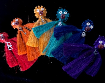 Op de afbeelding: Zes kleurrijke garenpoppen met verschillende bedels die aan hun nek hangen. De poppen zijn gerangschikt in een regenboogpatroon van rood tot paars.