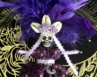 May include: A doll featuring a skull head adorned with a crown of white cowrie shells and small lavender fabric roses. The doll wears a dark purple dress with metallic copper polka dots, a silver crescent moon charm, and a large purple feather headdress.