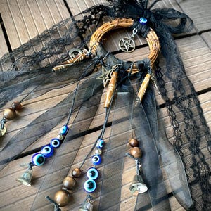 May include: A decorative wall hanging featuring a circular wooden hoop adorned with black lace ribbons, a silver pentagram charm, a star charm, and a crescent moon charm. The piece includes dangling strands with blue evil eye beads, brown wooden beads, and small silver-toned metal bells.
