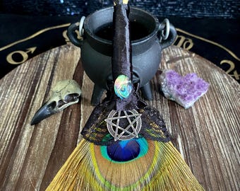 May include: A decorative broom featuring a handle wrapped in black satin ribbon, adorned with an iridescent abalone cabochon and a silver-toned metal pentagram charm. The bristles are made of a vibrant peacock feather with black lace trim.