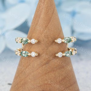 Peut inclure: Deux bagues en or, ornées de petits pierres précieuses rondes et en forme de marquise, sont présentées sur un cône en bois. Les bagues présentent un mélange de pierres claires et vertes, créant un effet scintillant. Les bagues sont posées sur un fond bleu doux.