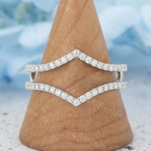May include: A pair of silver-toned, V-shaped rings, encrusted with small, clear gemstones. The rings are displayed on a wooden cone-shaped stand. The background is a soft blue with the text "AmandaFineJewelry".