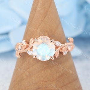 Könnte beinhalten: Verlobungsring aus Roségold mit einem ovalen, hellblauen Edelstein, der von kleineren Diamanten umgeben ist. Der Ring hat ein zartes florales Design mit Blättern und Ranken.