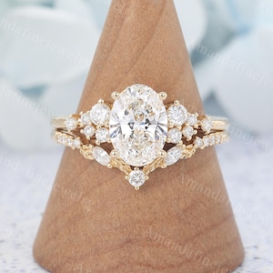 May include: A gold engagement ring set featuring a large oval-cut center stone surrounded by smaller round and marquise-cut accent diamonds. The band includes intricate leaf-like metal detailing and a curved design that nests with the main ring.