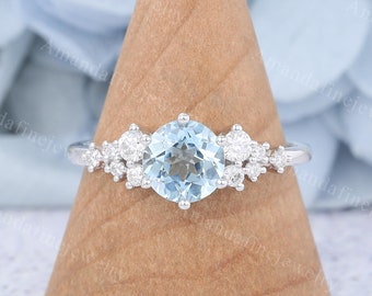 May include: A silver ring with a light blue gemstone surrounded by smaller clear gemstones. The ring is on a wooden ring holder.