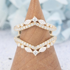 Pode incluir: Duas alianças de casamento douradas cravejadas de diamantes. Cada aliança tem um design em forma de V com uma fileira de pequenos diamantes e pedras em forma de marquesa. Os anéis são exibidos em um cone de madeira.