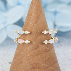 May include: Two gold rings with opal and diamond accents are displayed on a wooden cone. The rings feature oval opals and small round diamonds. The rings are set in a gold band. The background is a soft blue.