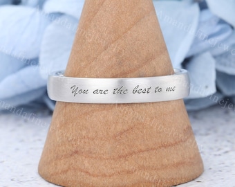 May include: A silver ring inscribed with "You are the best to me" is presented on a wooden cone. The ring features a flat band design and is set against a soft blue backdrop.