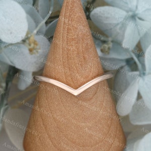 May include: A rose gold V-shaped ring displayed on a wooden cone. The ring has a simple, elegant design. The background features soft, blurred blue and white floral elements. The ring is a delicate piece of jewelry.