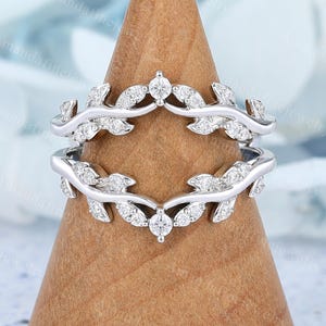 May include: A silver-toned ring guard featuring two bands connected by a leaf-inspired design. Each leaf segment is accented with small, clear, round-cut stones. The metal has a polished finish, and the structure is designed to frame a center stone.