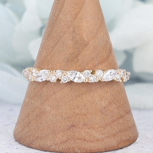 Può includere: Un anello a fascia in oro con una serie alternata di pietre preziose rotonde e a forma di marquise. L'anello è esposto su un supporto a forma di cono in legno, che mette in risalto il suo design intricato e la sua brillantezza. L'anello è un gioiello.