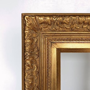 May include: A close-up of a gold-coloured, ornate picture frame. The frame has detailed carvings and a textured surface. The design is classic and elegant, with a rectangular opening for a picture or artwork.