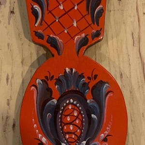 May include: A hand-painted wooden decorative spoon with a vibrant red background. The spoon features intricate floral and geometric designs in blue, white, and black. The spoon is approximately 10 inches long.