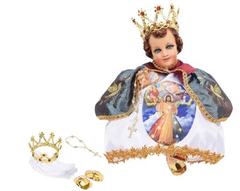 May include: A figurine of the Child Jesus wearing a white robe with a gold trim, a blue and green cape, and a gold crown. The figurine is holding a rosary and has a depiction of the Divine Mercy image on the front of the robe.
