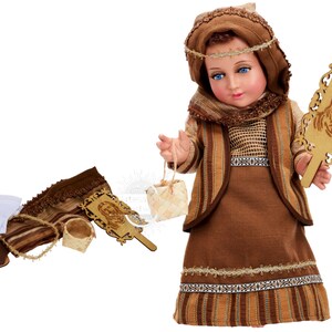 May include: A brown and beige doll dressed in a traditional outfit with a brown head scarf, holding a wooden frame with a carved image of Jesus. The doll is part of a nativity set and includes accessories like a basket, sandals, and a cloth.