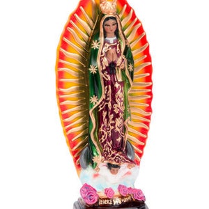 May include: A figurine of the Virgin Mary, Our Lady of Guadalupe, standing on a cloud with a crown and a blue robe over a burgundy dress. She is holding her hands in prayer. The figurine is surrounded by a golden halo with red and yellow rays. The base of the figurine is brown and has the words "REINO DE DIOS" written on it.