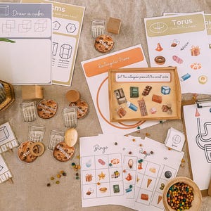 Op de afbeelding: Educatief materiaal voor kinderen, waaronder kaarten met geometrische vormen, een houten dienblad met objecten en bingokaarten. De afbeelding toont tekeningen van kubussen, rechthoekige prisma's en andere vormen, samen met kleine houten blokken en heldere glazen.