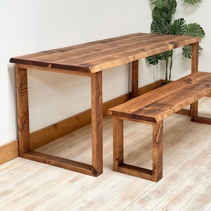 May include: A wooden dining table and bench set. The table has a rectangular top and square legs. The bench is shorter and has a similar design. Both pieces are made of stained wood with a natural grain.