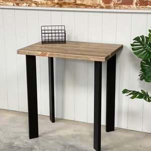 May include: A rustic wooden desk with black metal legs. The desk has a natural wood finish and is supported by four black metal legs. The desk is sitting on a concrete floor in front of a brick wall.